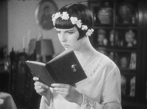 Animated gif of the silent film actress Louise Brooks reading a book, wearing a crown of flowers, and looking askance.